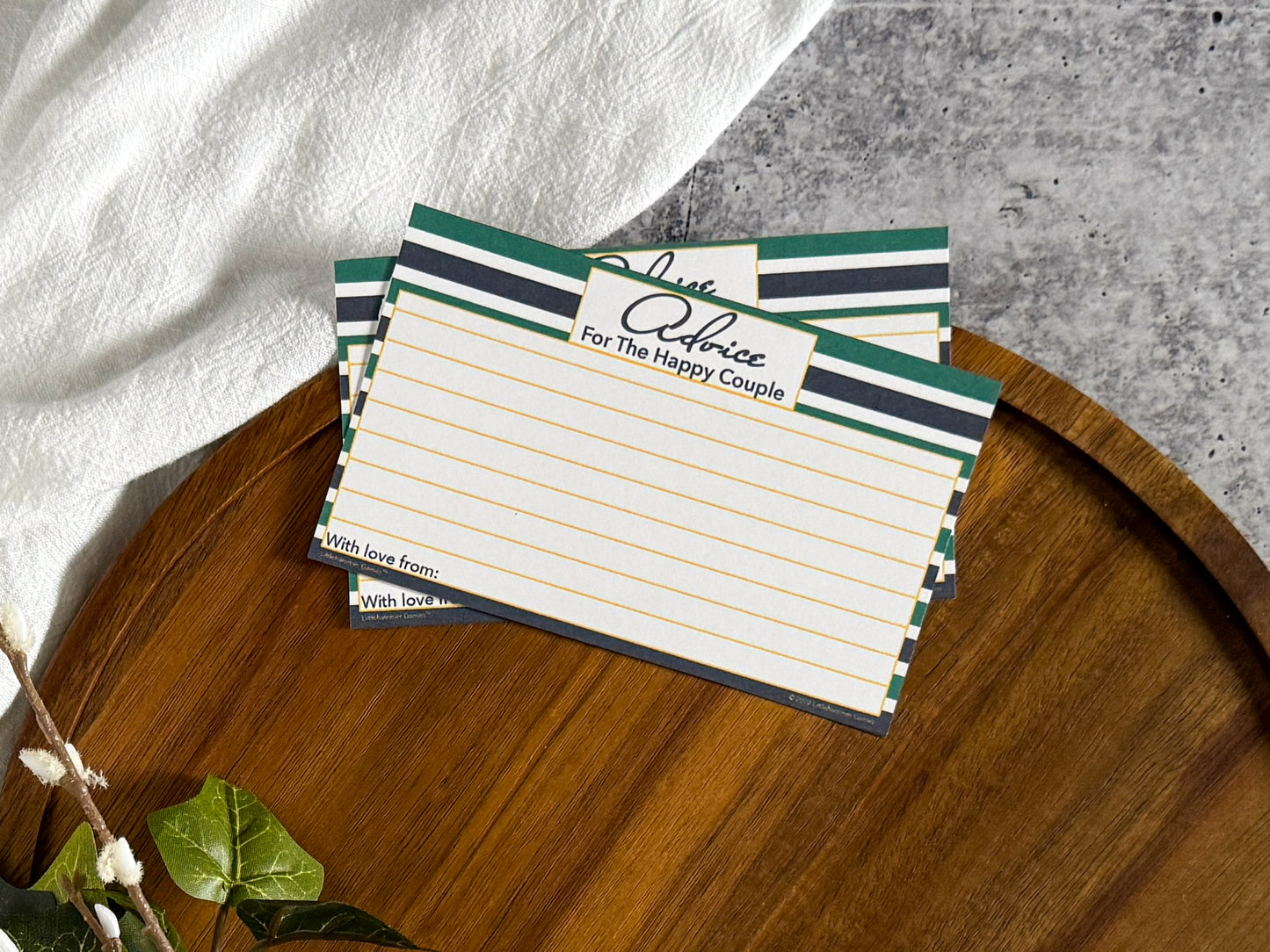 2 Green & Navy Stripe Advice for the Happy Couple cards laying on the top edge of a round wooden tray with a cream cloth and foliage surrounding the tray