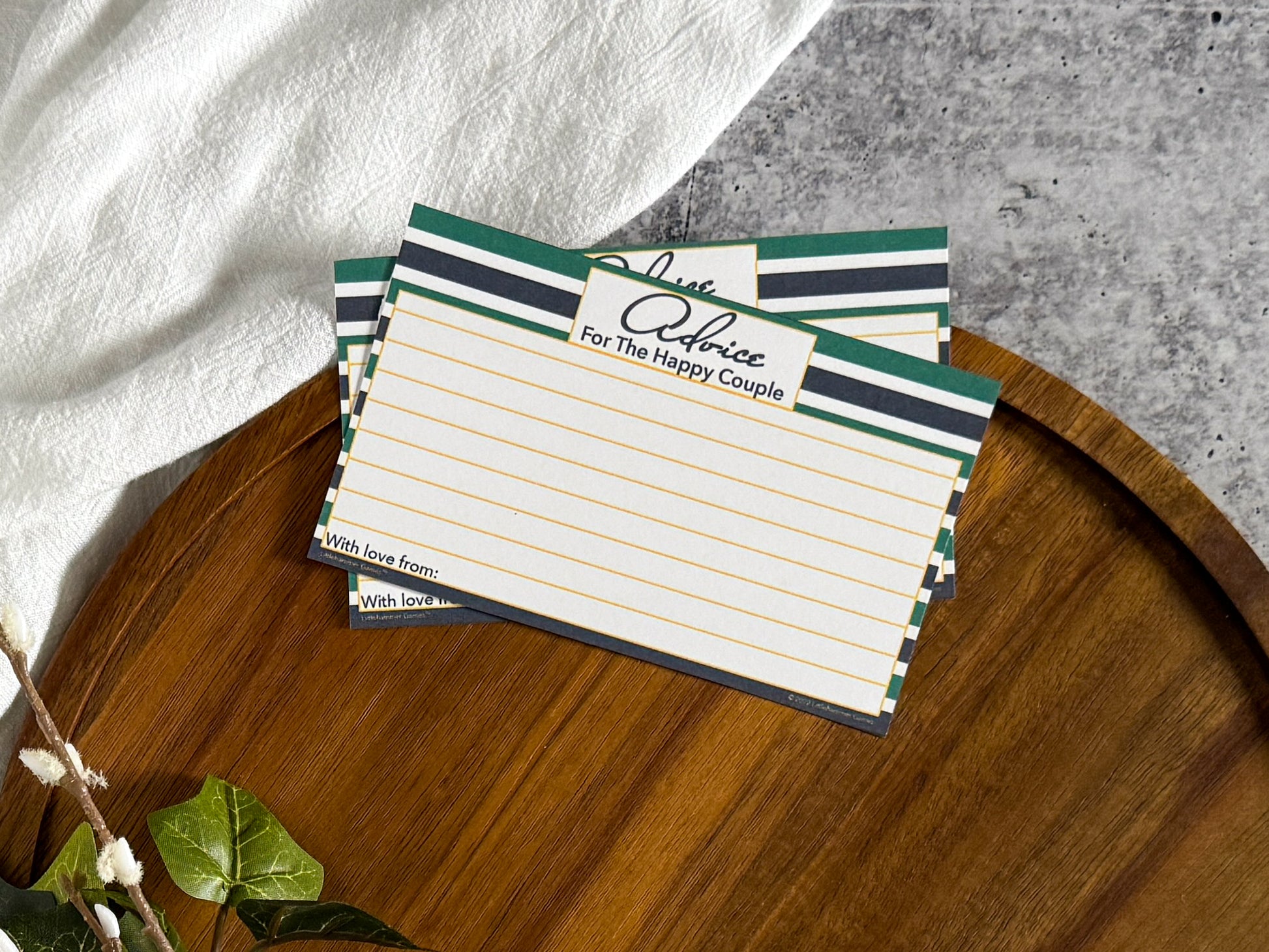 2 Green & Navy Stripe Advice for the Happy Couple cards laying on the top edge of a round wooden tray with a cream cloth and foliage surrounding the tray