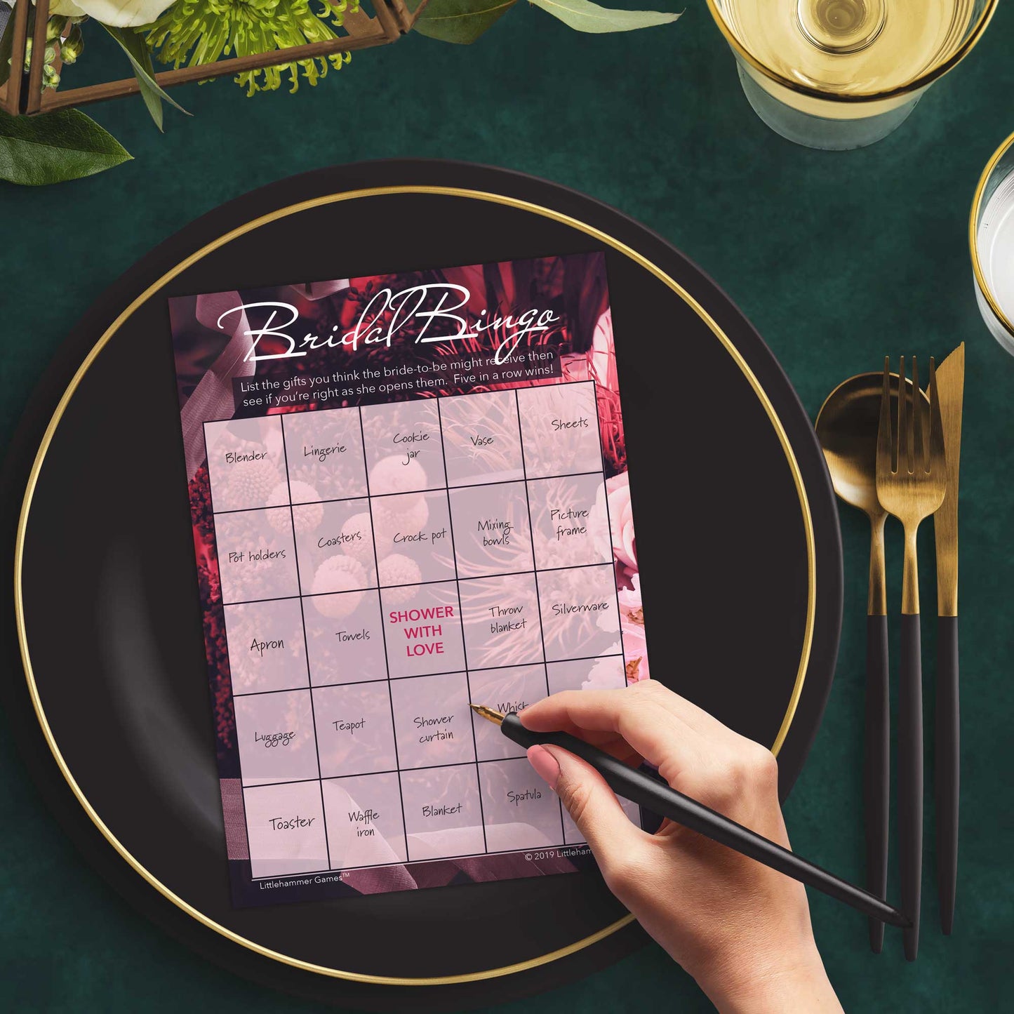 Woman with a pen sitting at a dark place setting with a black and gold plate filling out a floral-themed Bridal Gift Bingo card