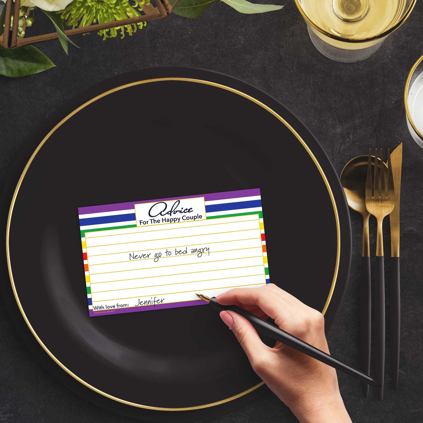 Woman with a pen sitting at a dark place setting with a black and gold plate filling out a rainbow-striped Advice for the Happy Couple card