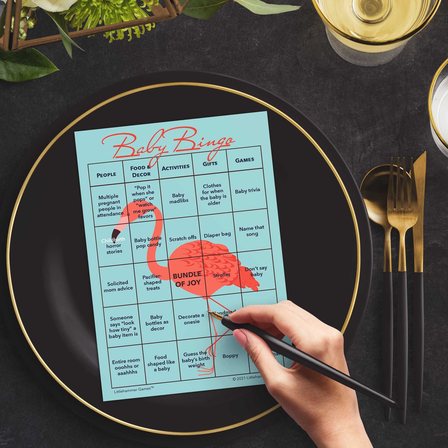 Woman with a pen sitting at a table with a flamingo-themed Baby Bingo game card on a black and gold plate at a dark place setting