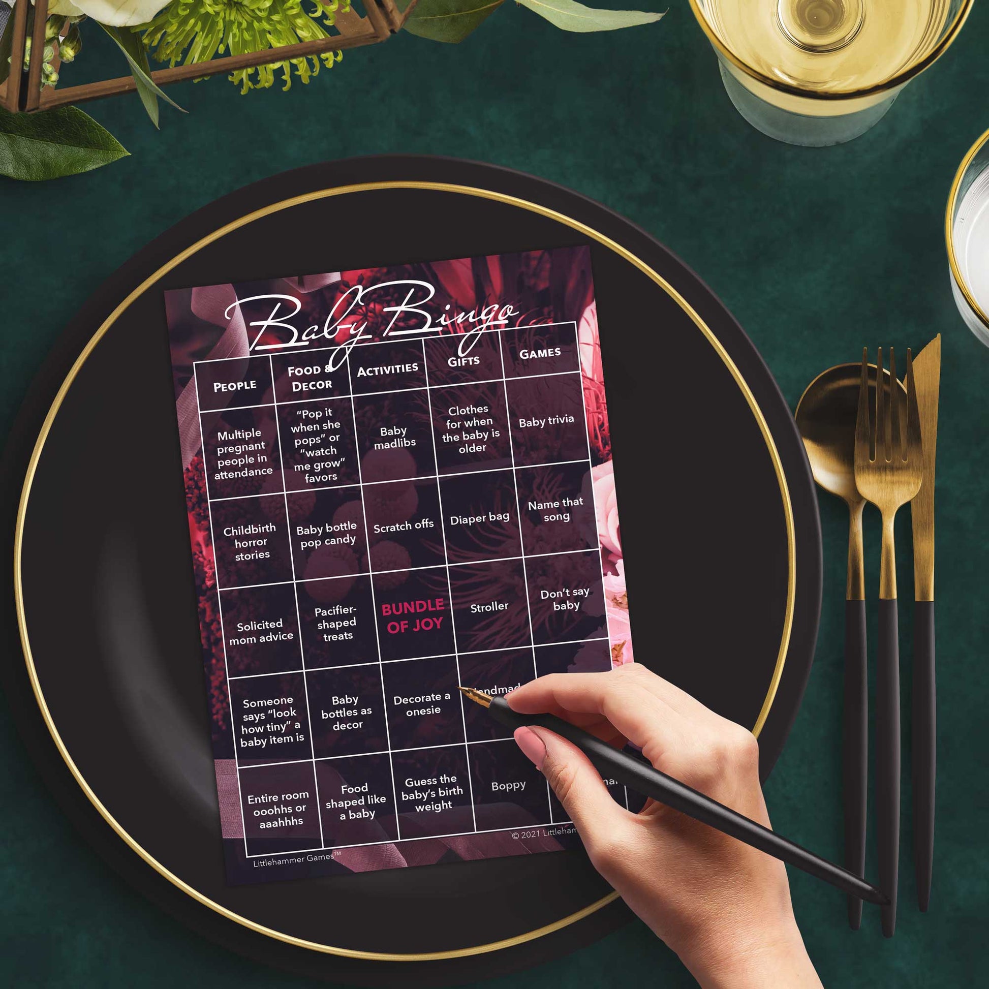Woman with a pen sitting at a table with a fall leaves Baby Bingo game card on a black and gold plate at a dark place setting