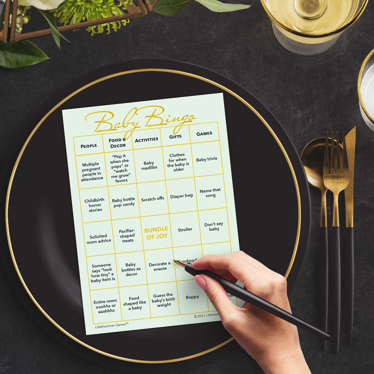 Woman with a pen sitting at a table with a mint and gold Baby Bingo game card on a black and gold plate at a dark place setting