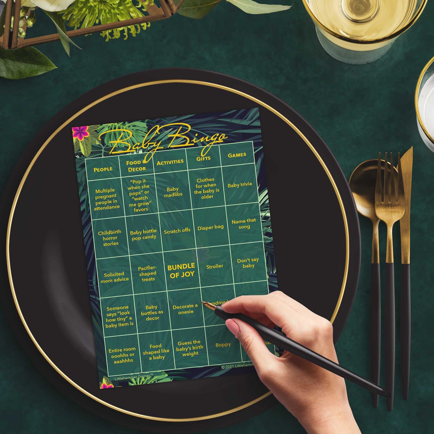 Woman with a pen sitting at a table with a tropical-themed Baby Bingo game card on a black and gold plate at a dark place setting