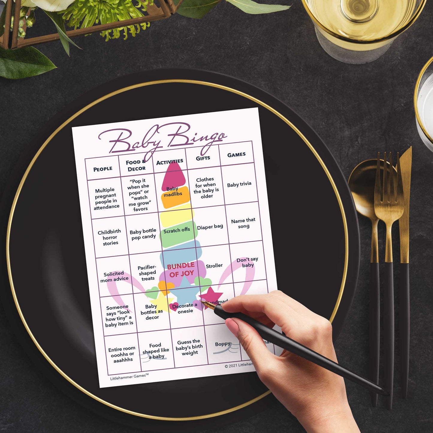 Woman with a pen sitting at a table with a unicorn-themed Baby Bingo game card on a black and gold plate at a dark place setting