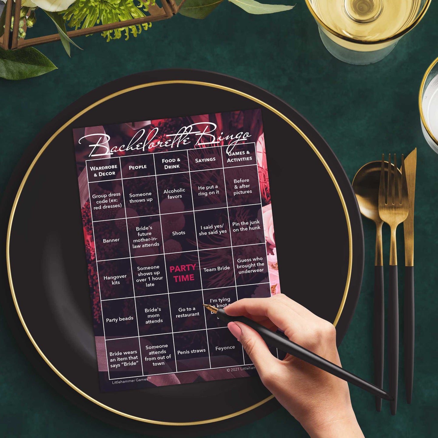Woman with a pen sitting at a table with a dark floral themed Bachelorette Bingo game card on a gold and black plate on a dark place setting