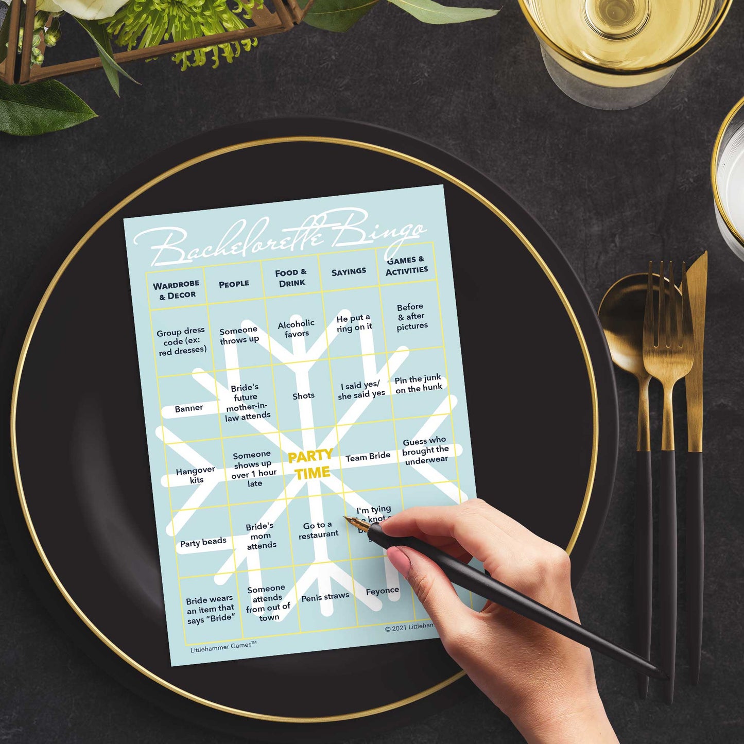 Woman with a pen sitting at a table with a snowflake themed Bachelorette Bingo game card on a gold and black plate on a dark place setting