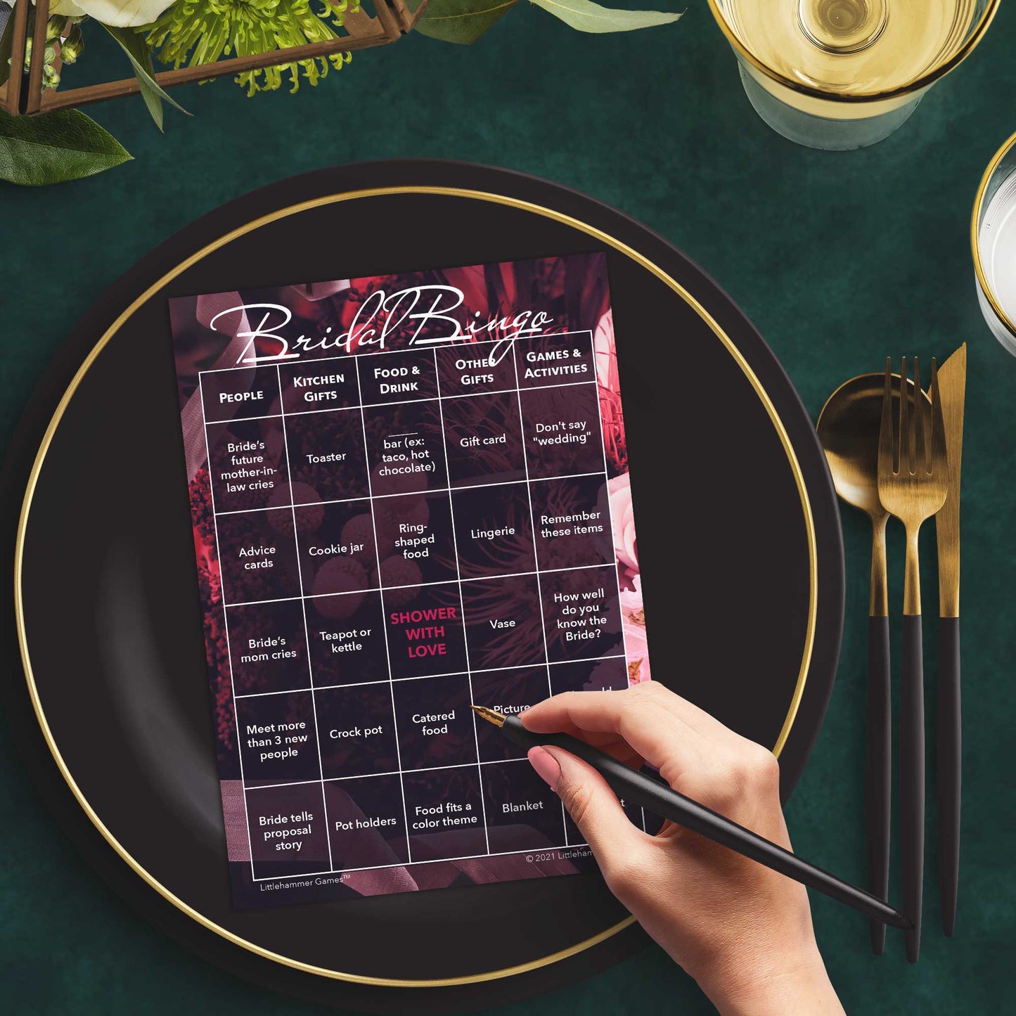 Woman with a pen sitting at a table with a dark floral themed Bridal Bingo game card on a black and gold plate at a dark place setting