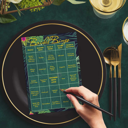 Woman with a pen sitting at a table with a tropical-themed Bridal Bingo game card on a black and gold plate at a dark place setting