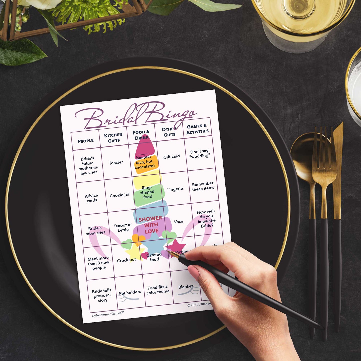 Woman with a pen sitting at a table with a unicorn-themed Bridal Bingo game card on a black and gold plate at a dark place setting