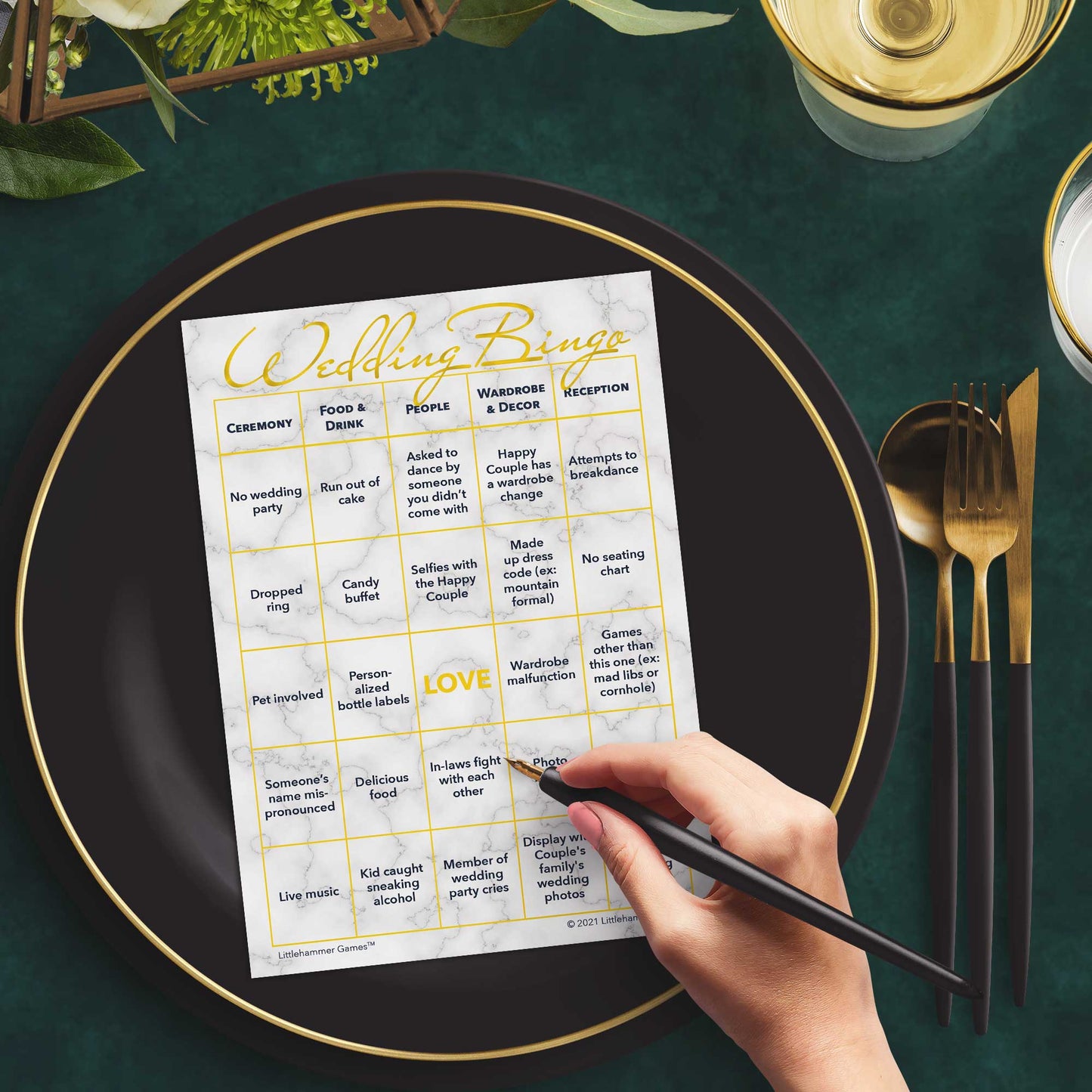 Woman with a pen sitting at a table with a gold and marble Wedding Bingo game card on a black and gold plate at a dark place setting