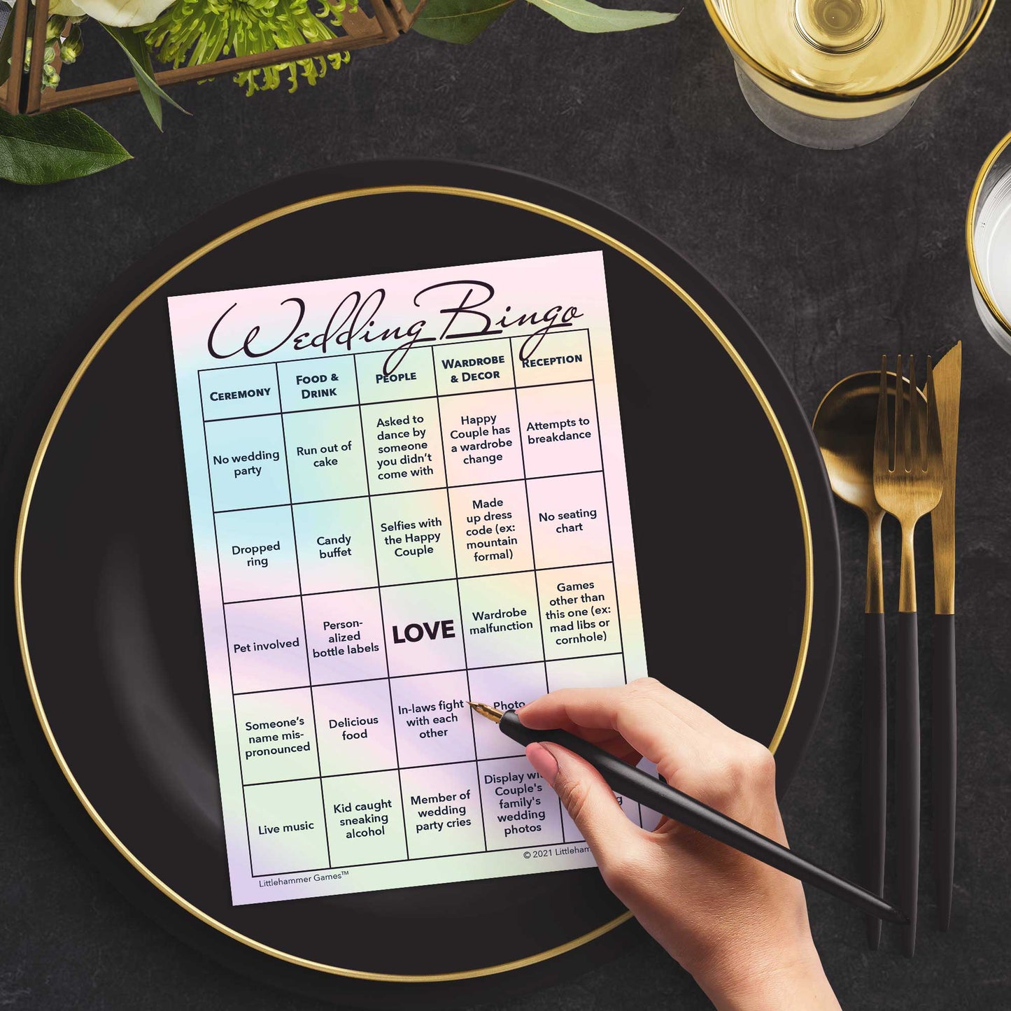 Woman with a pen sitting at a table with a hologram themed Wedding Bingo game card on a black and gold plate at a dark place setting