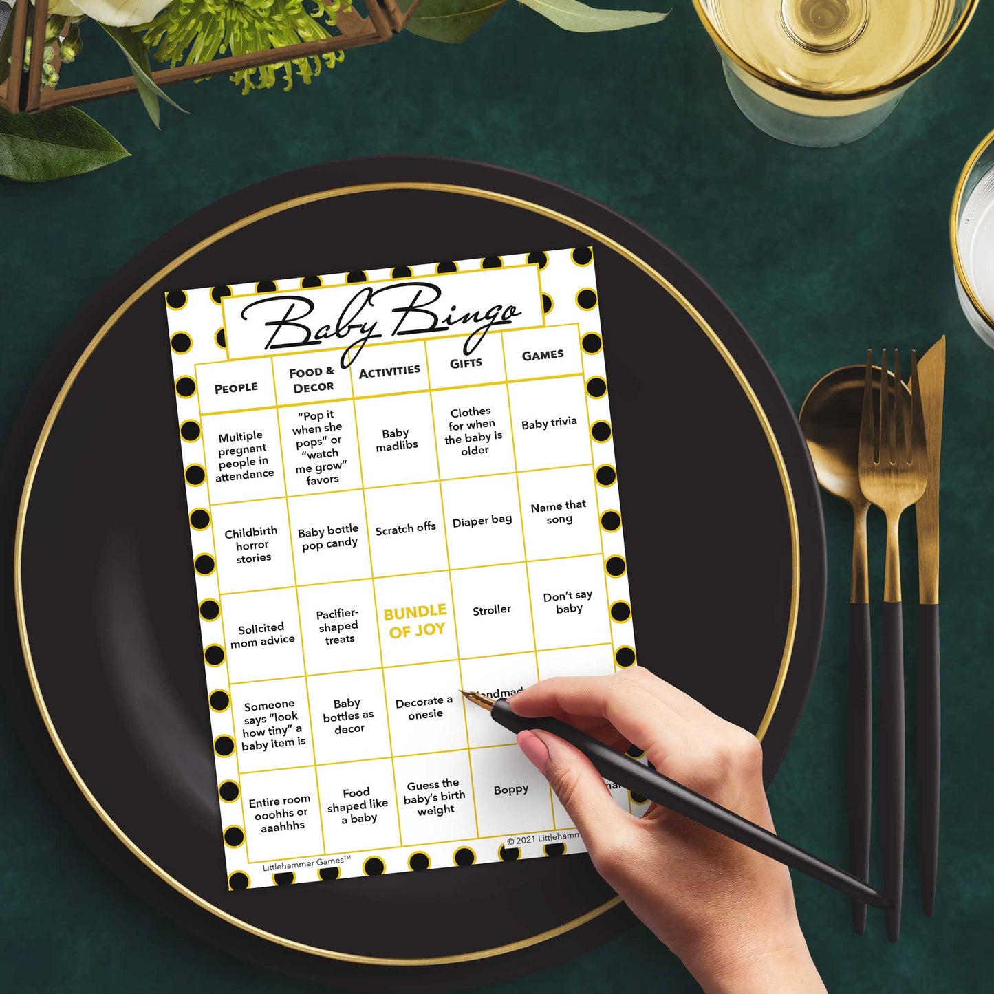 Woman with a pen sitting at a table with a black and gold polka dot Baby Bingo game card on a black and gold plate at a dark place setting