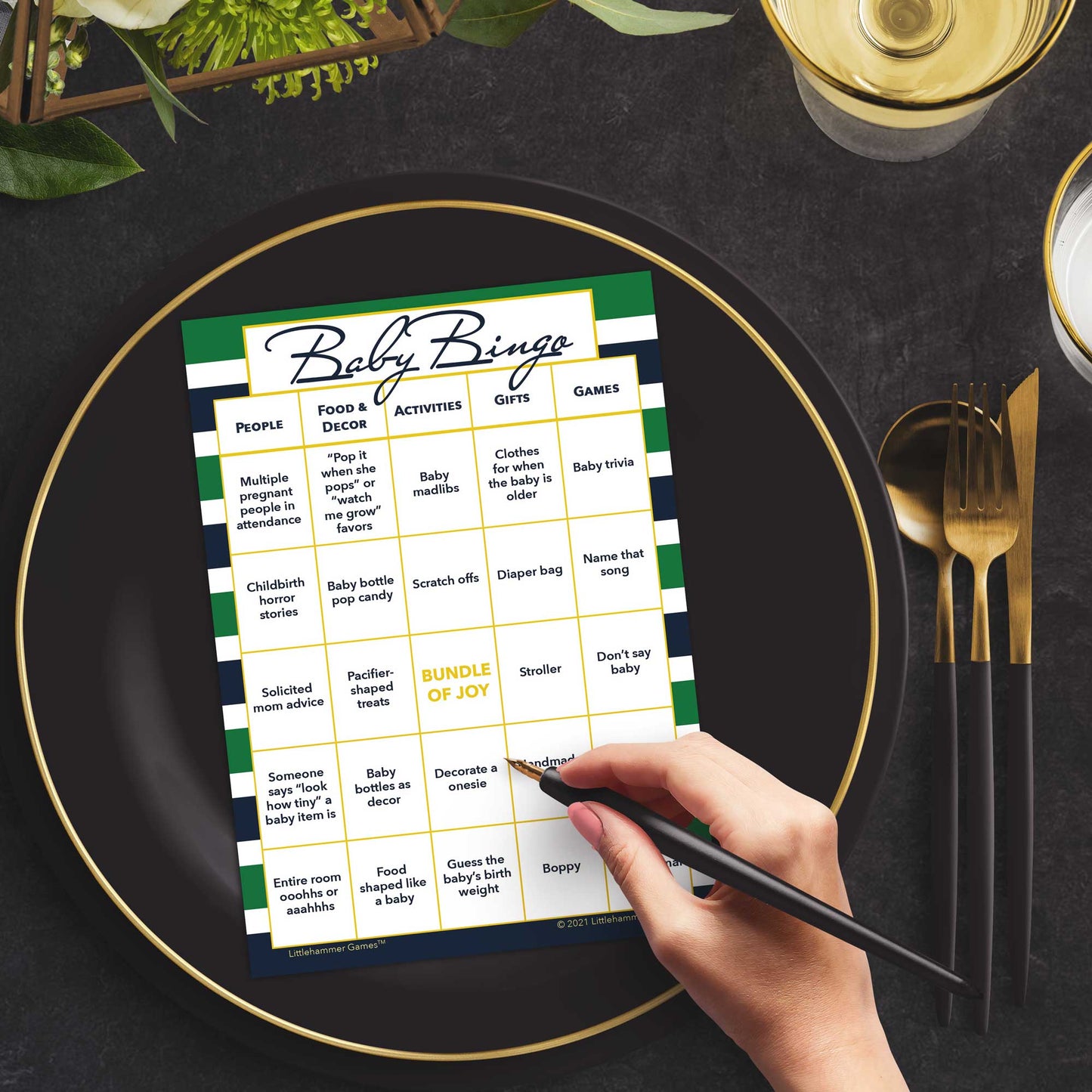 Woman with a pen sitting at a table with a green and navy-striped Baby Bingo game card on a black and gold plate at a dark place setting