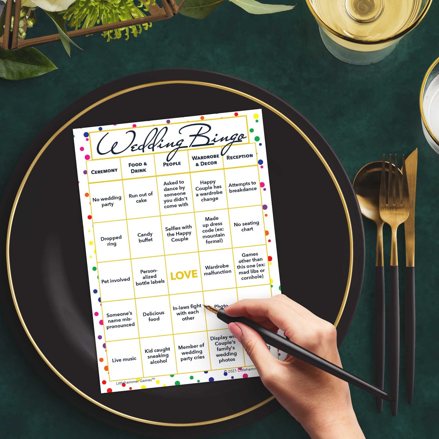 Woman with a pen sitting at a table with a rainbow polka dot Wedding Bingo game card on a black and gold plate at a dark place setting