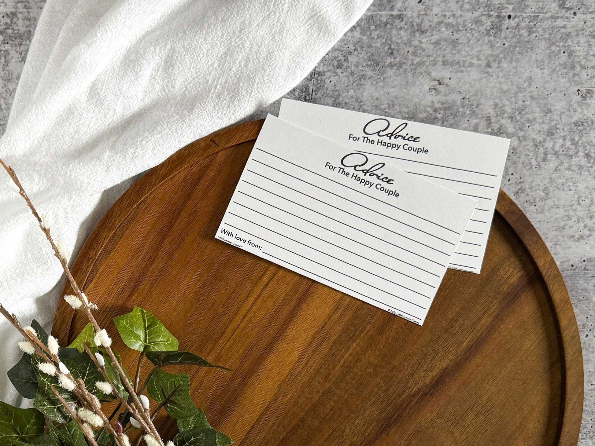 2 Black & White Advice for the Bachelorette cards laying on the top edge of a round wooden tray with a cream cloth and foliage surrounding the tray