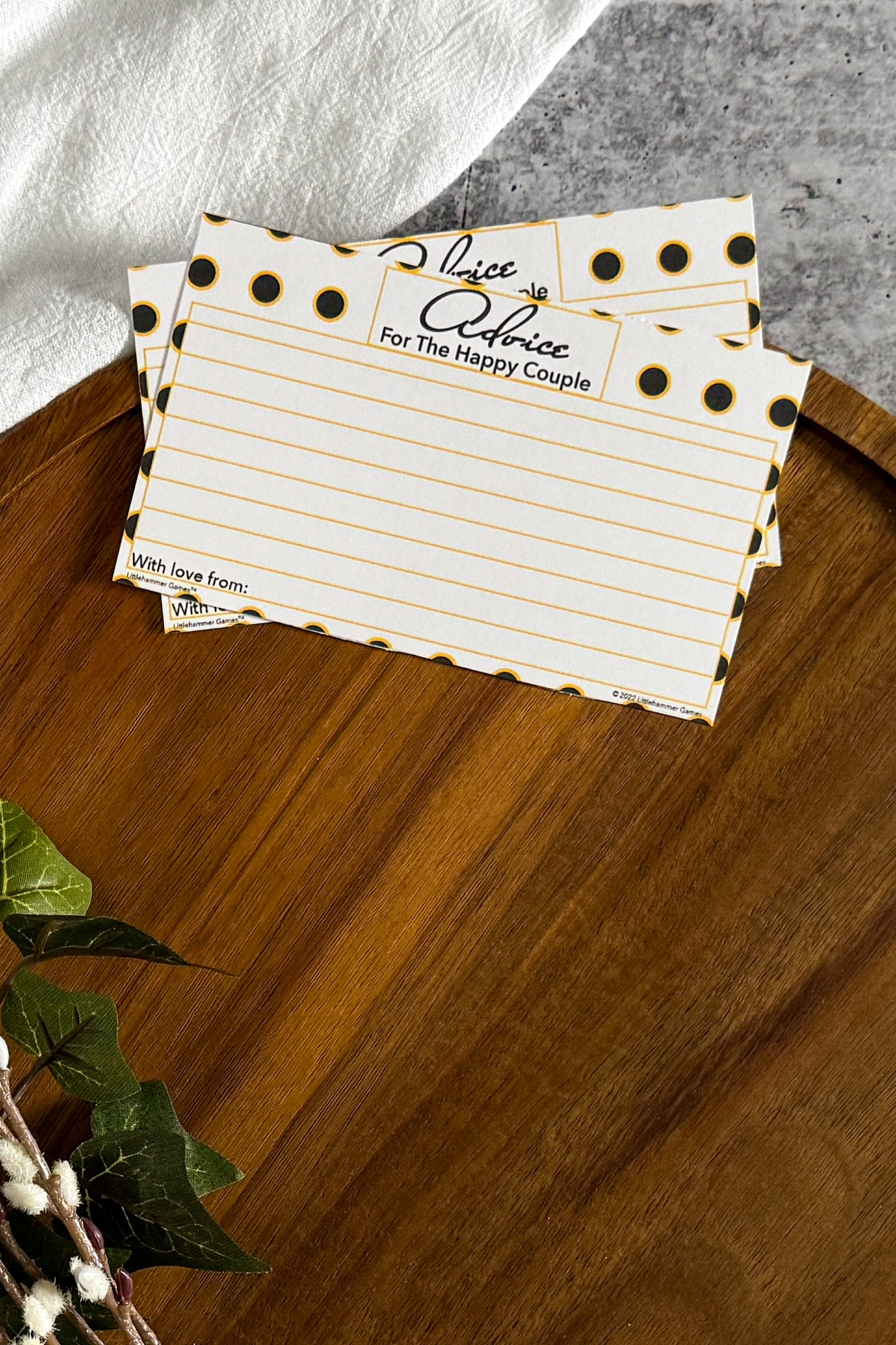 2 Black & Gold Polka Dot Advice for the Happy Couple cards laying on the top edge of a round wooden tray with a cream cloth and foliage surrounding the tray