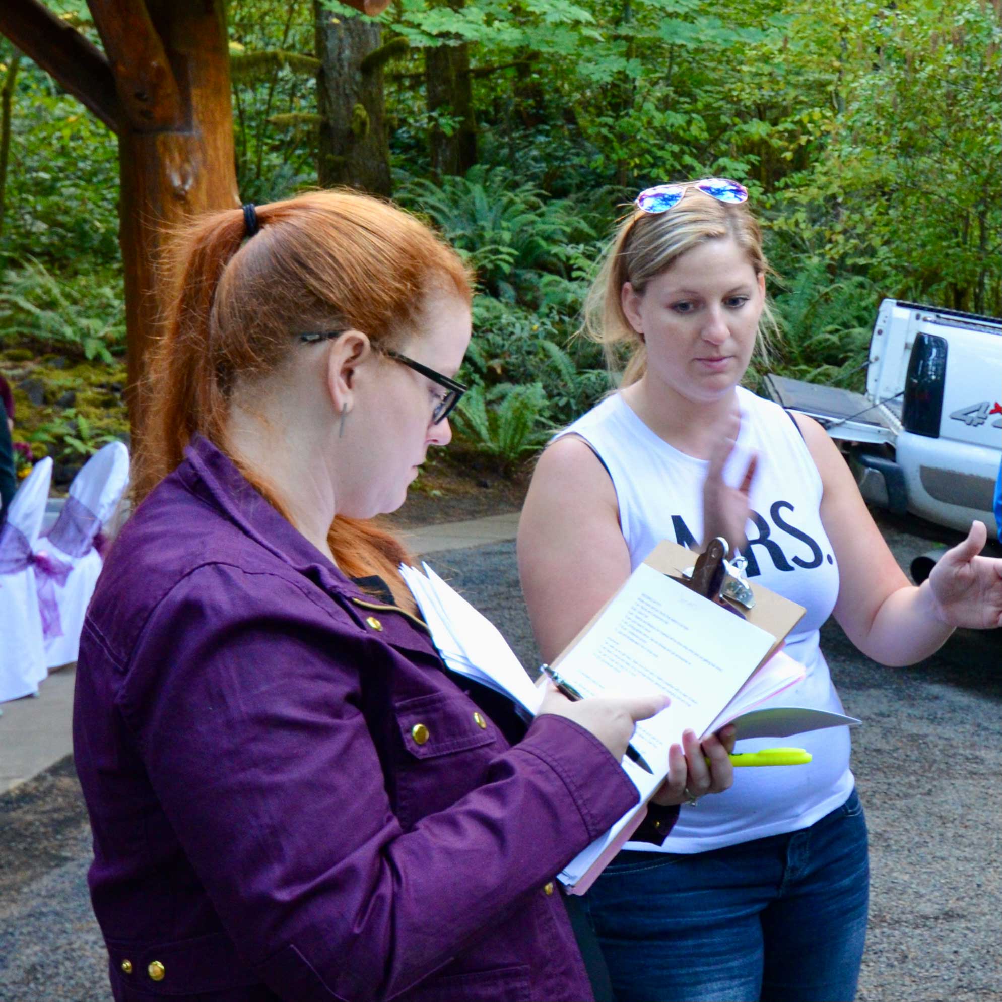 Woman with a white tank top that says "Mrs" talking to a woman with red hair in a ponytail wearing a purple jacket and holding a clipboard