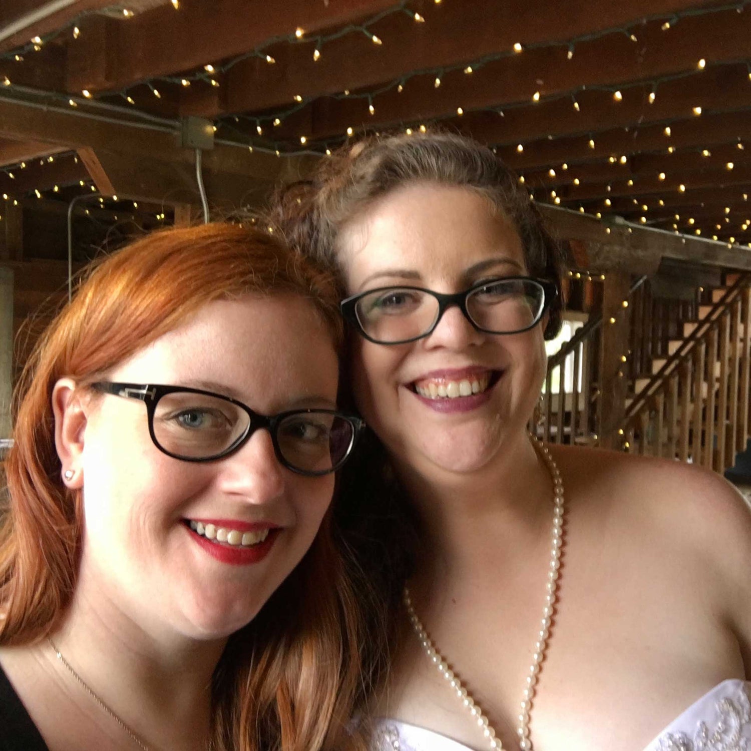 Red haired woman with glasses next to a bride with brown hair and glasses wearing a wedding dress and pearl necklace