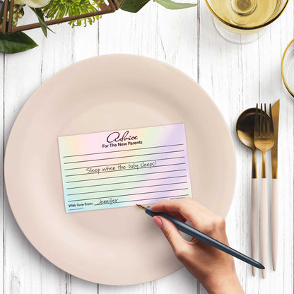 Woman’s hand writing on a hologram-themed baby shower advice card at a light place setting