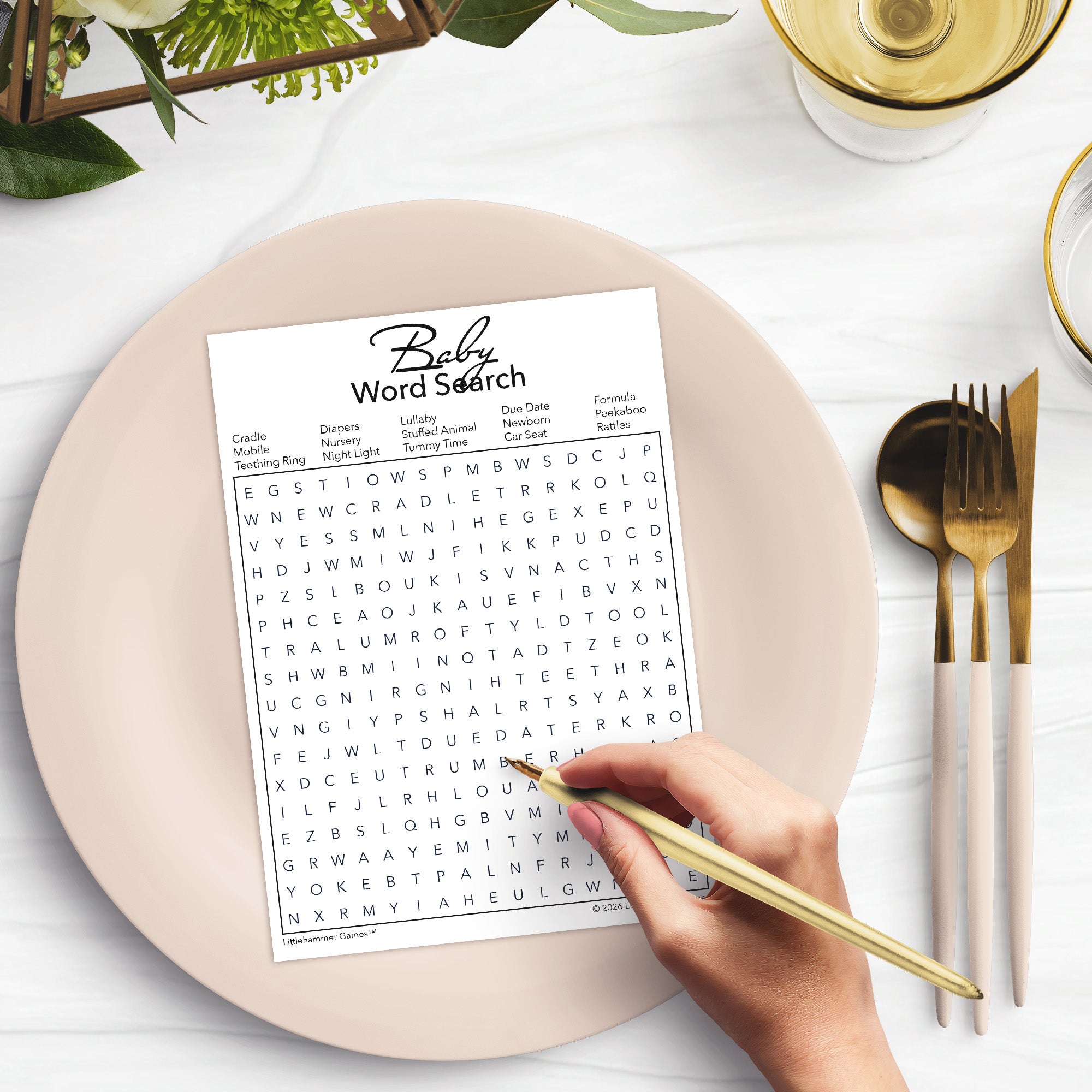 Hand holding a gold pen over a printable baby word search game card on a pale pink plate with utensils and decor.