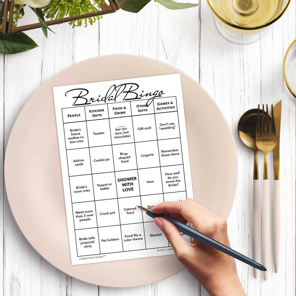 Woman’s hand holding a pen playing a black and white Bridal Bingo game card at a light place setting