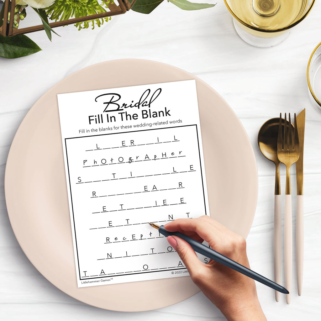 Woman’s hand holding a pen playing a black and white Bridal Fill in the Blank game card at a light place setting