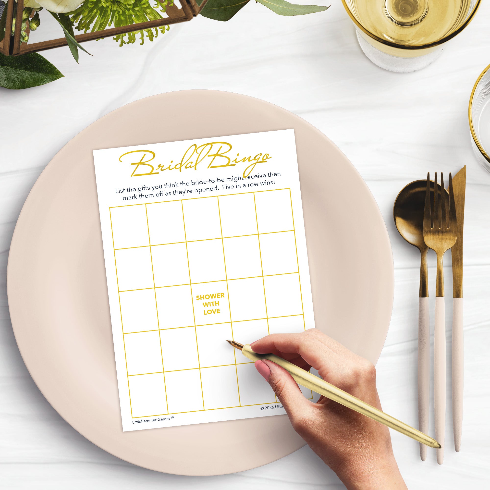 Woman’s hand holding a pen playing a gold and white Bridal Gift Bingo game card at a light place setting