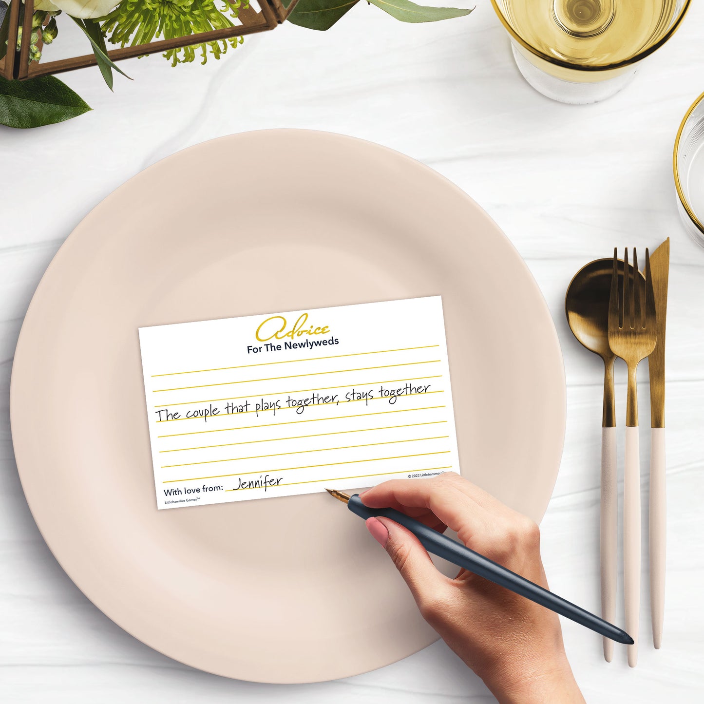 Woman’s hand holding a pen writing a note on a gold Advice for the Newlyweds card at a light place setting