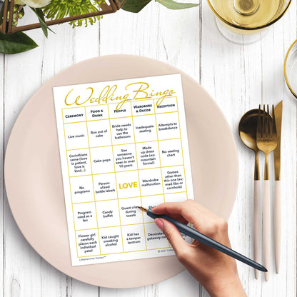 Woman’s hand holding a pen playing a gold and white Bride and Groom Wedding Bingo game card at a light place setting