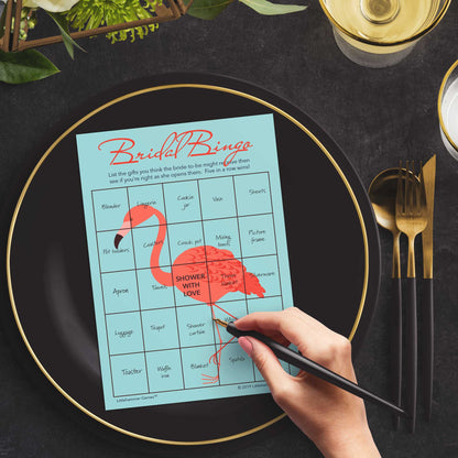 Woman with a pen sitting at a dark place setting with a black and gold plate filling out a flamingo-themed Bridal Gift Bingo card