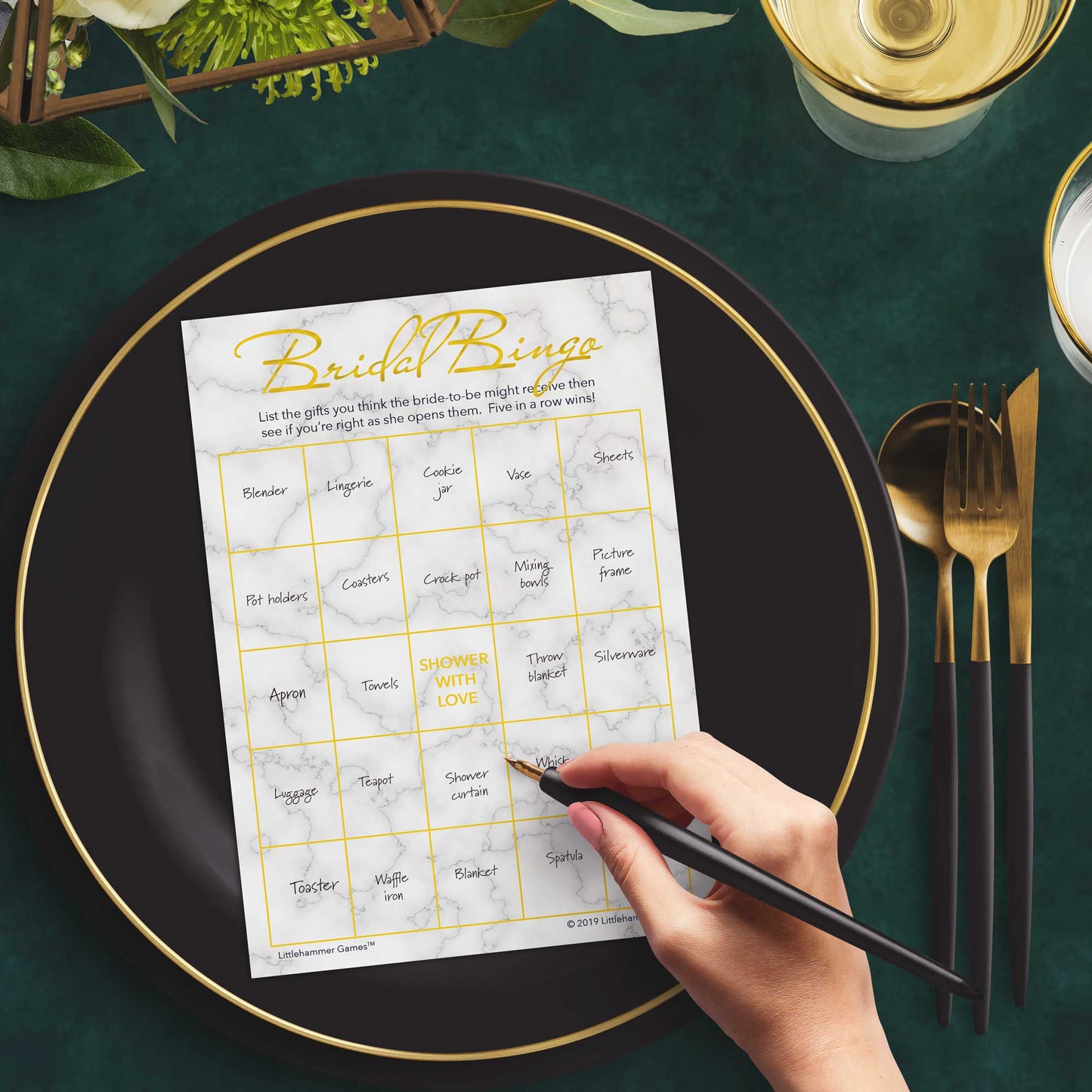 Woman with a pen sitting at a dark place setting with a black and gold plate filling out a gold and marble Bridal Gift Bingo card