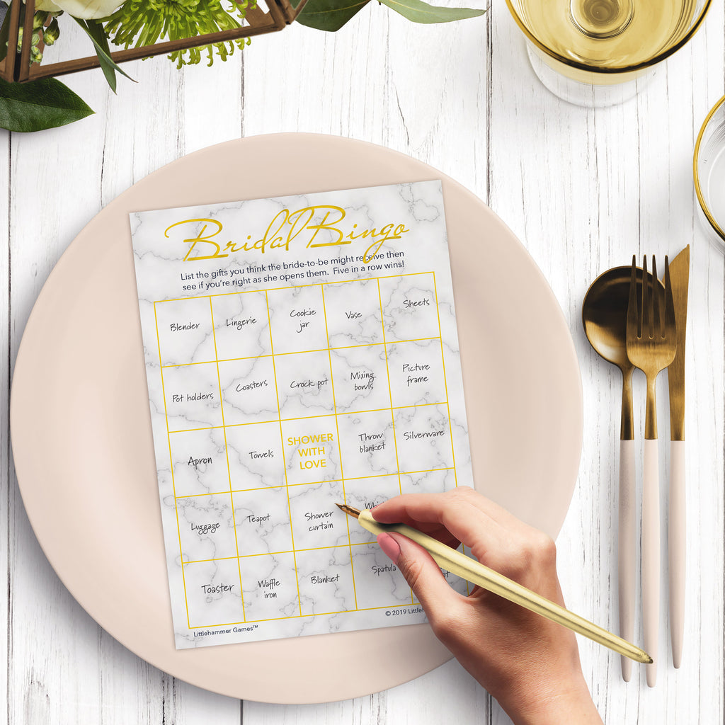 Woman with a pen sitting at a dark place setting with a black and gold plate filling out a gold and marble Bridal Gift Bingo card