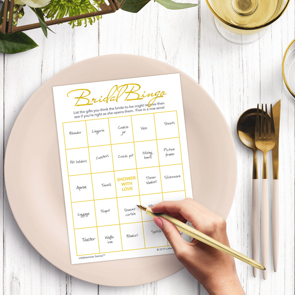 Woman’s hand holding a pen playing a gold and white Bridal Gift Bingo game card at a light place setting