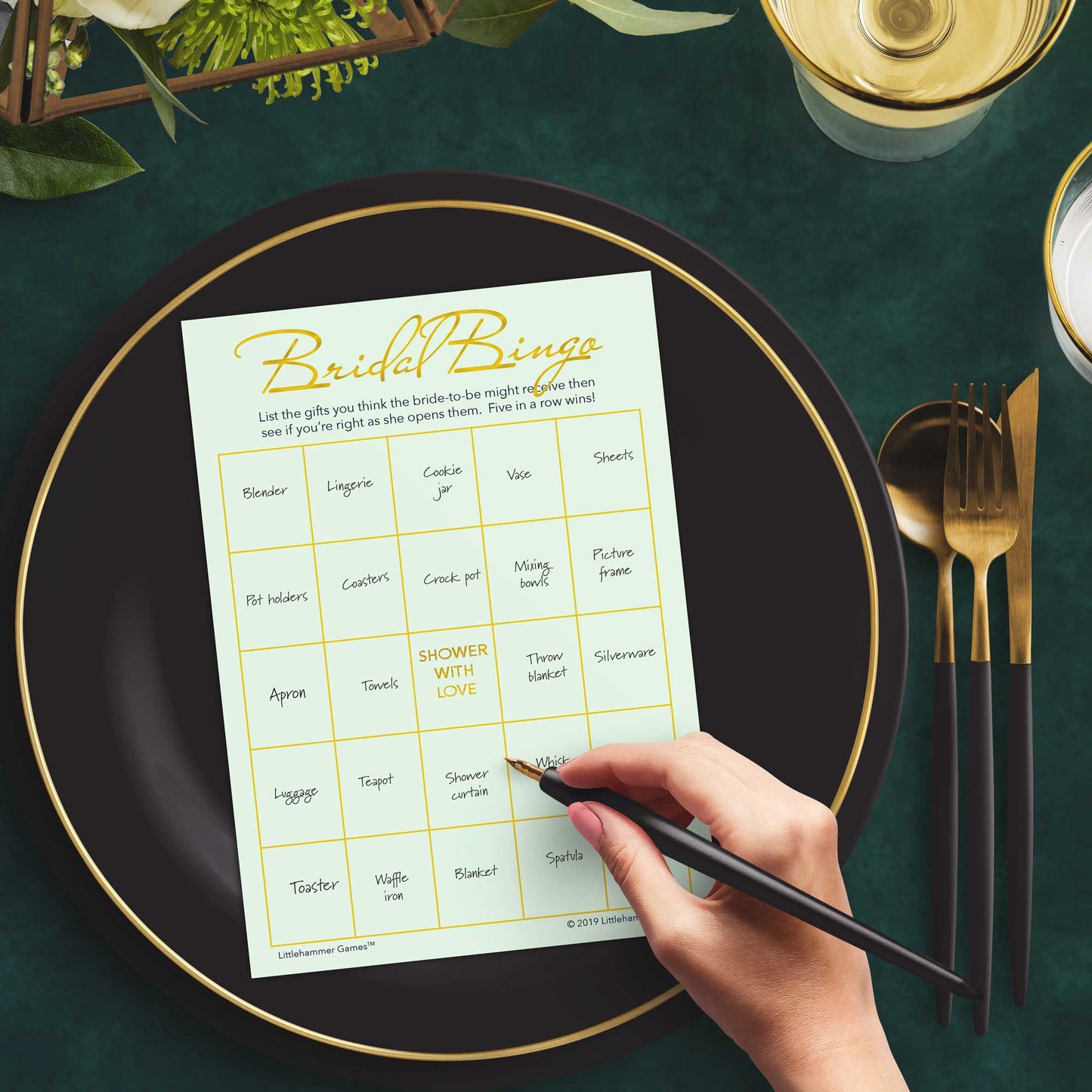 Woman with a pen sitting at a dark place setting with a black and gold plate filling out a mint and gold Bridal Gift Bingo card