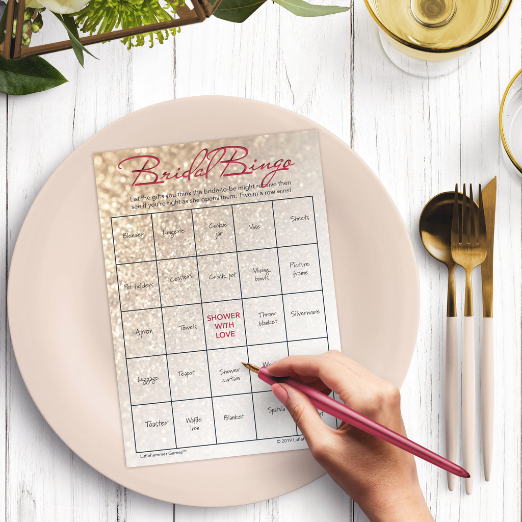 Woman with a pen sitting at a dark place setting with a black and gold plate filling out a glittery rose gold Bridal Gift Bingo card