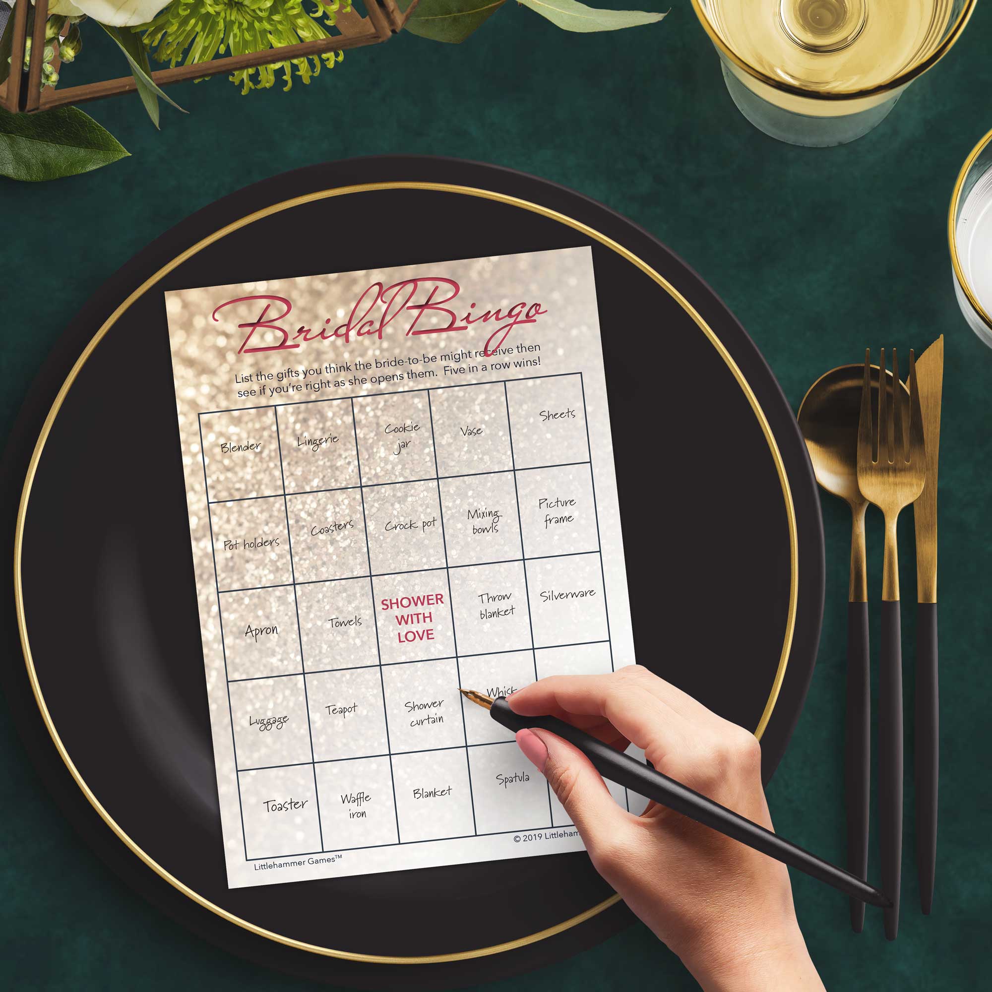 Woman with a pen sitting at a dark place setting with a black and gold plate filling out a glittery rose gold Bridal Gift Bingo card