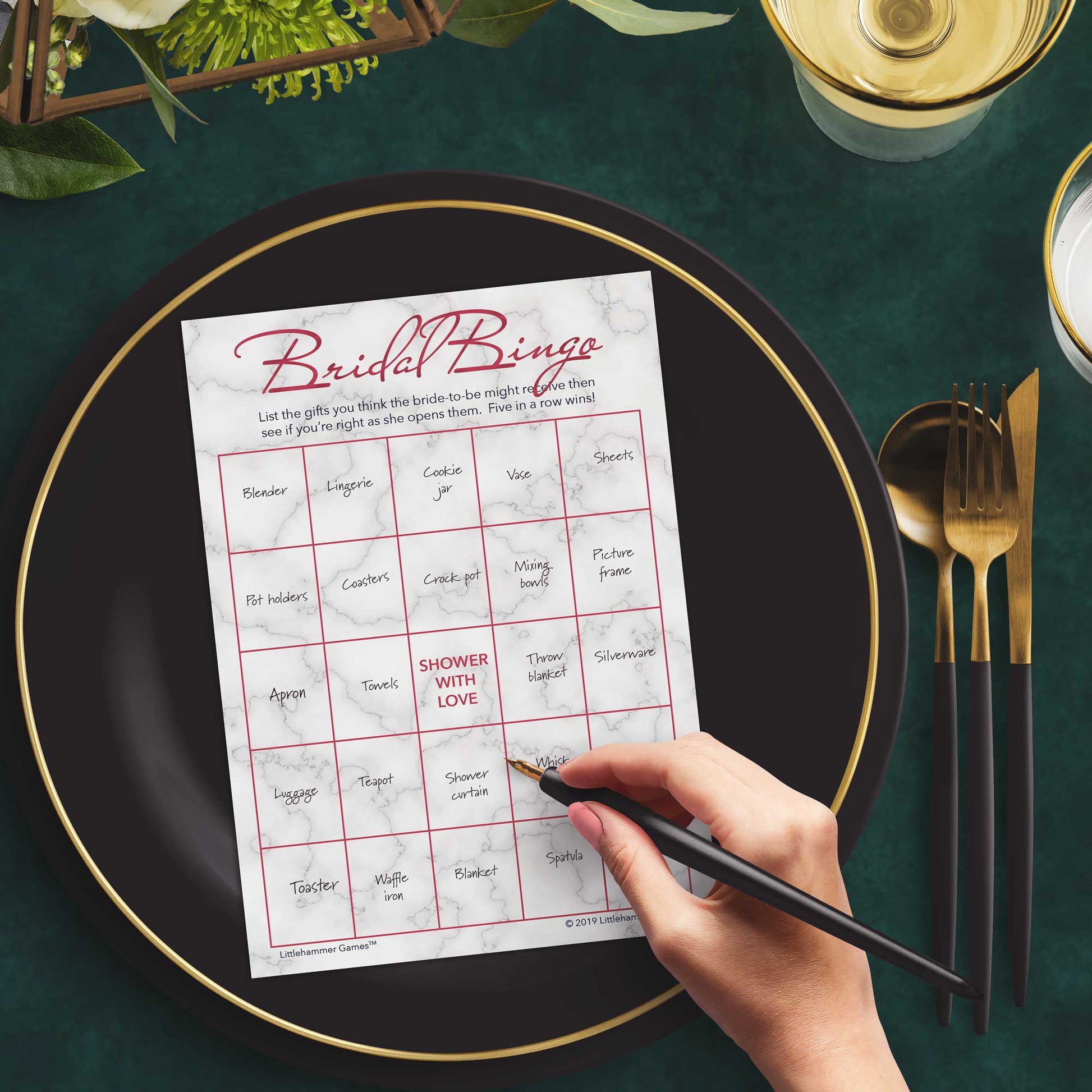 Woman with a pen sitting at a dark place setting with a black and gold plate filling out a rose gold and marble Bridal Gift Bingo card