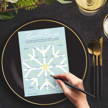 Woman with a pen sitting at a dark place setting with a black and gold plate filling out a snowflake-themed Bridal Gift Bingo card