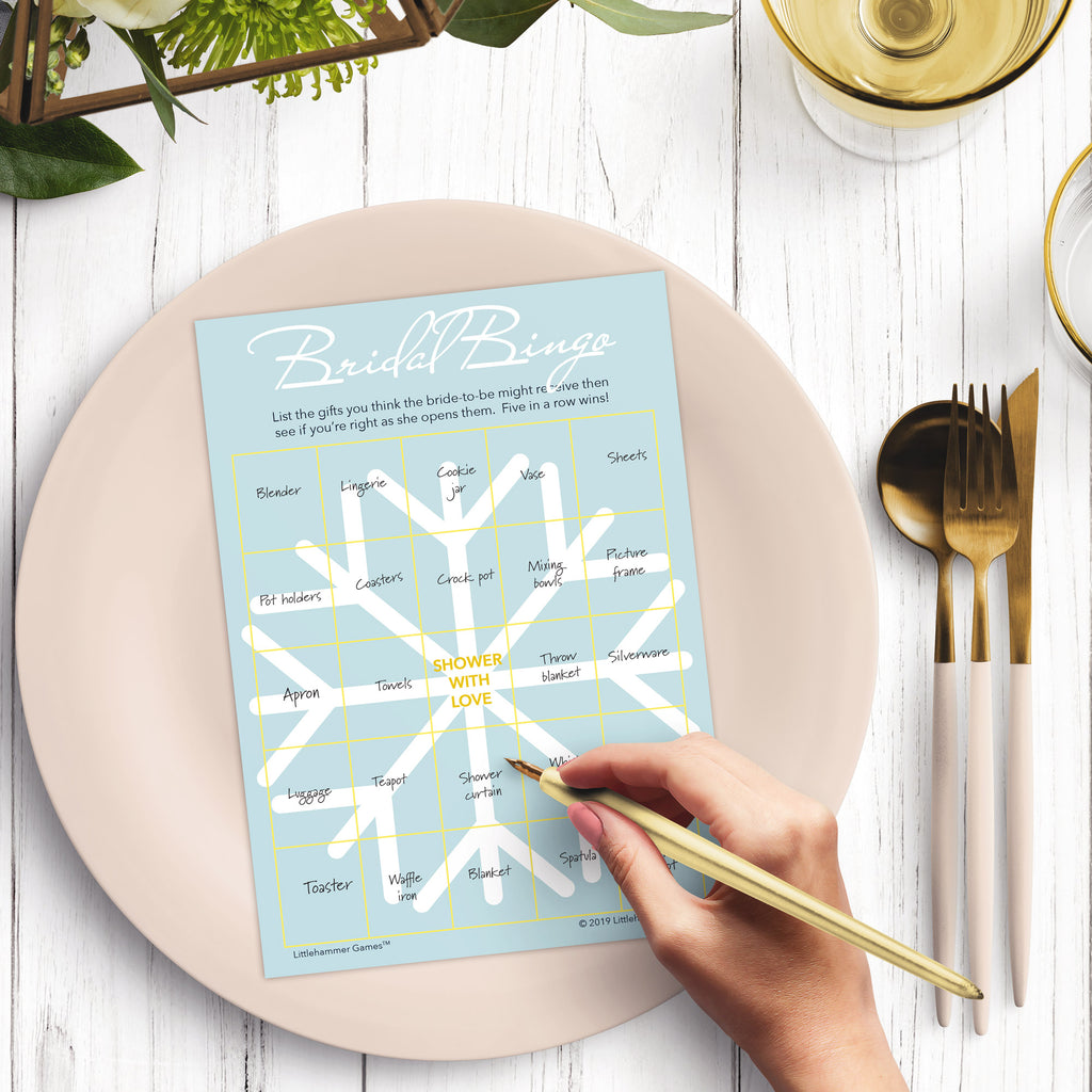 Woman with a pen sitting at a dark place setting with a black and gold plate filling out a snowflake-themed Bridal Gift Bingo card