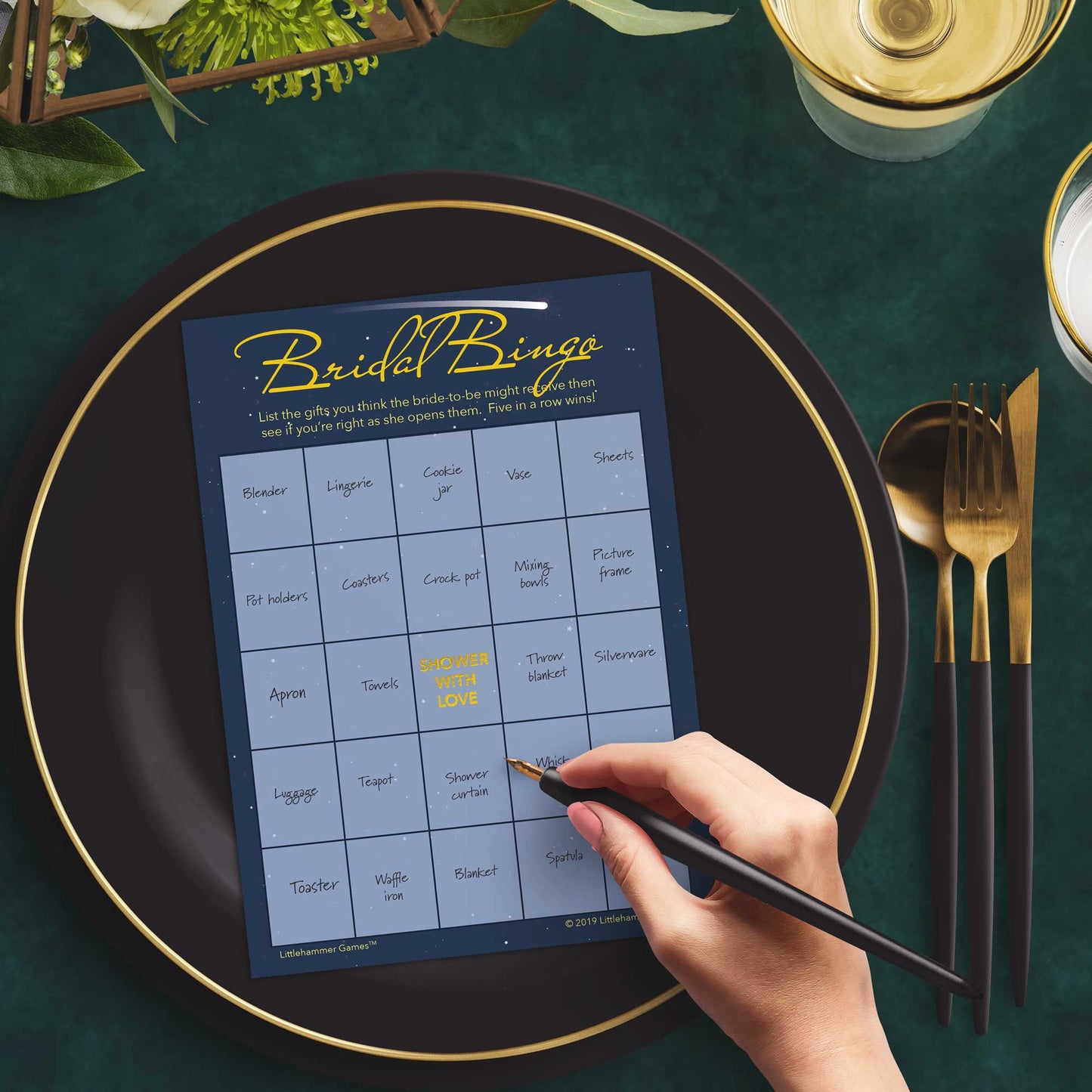 Woman with a pen sitting at a dark place setting with a black and gold plate filling out a celestial-themed Bridal Gift Bingo card