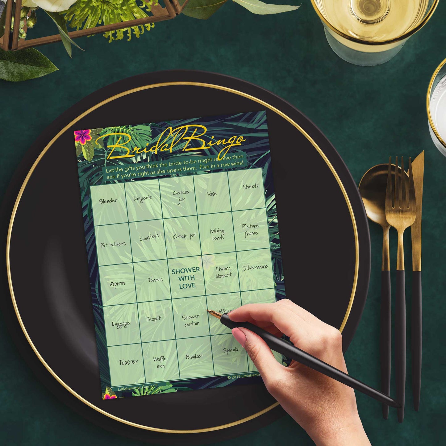 Woman with a pen sitting at a dark place setting with a black and gold plate filling out a tropical-themed Bridal Gift Bingo card