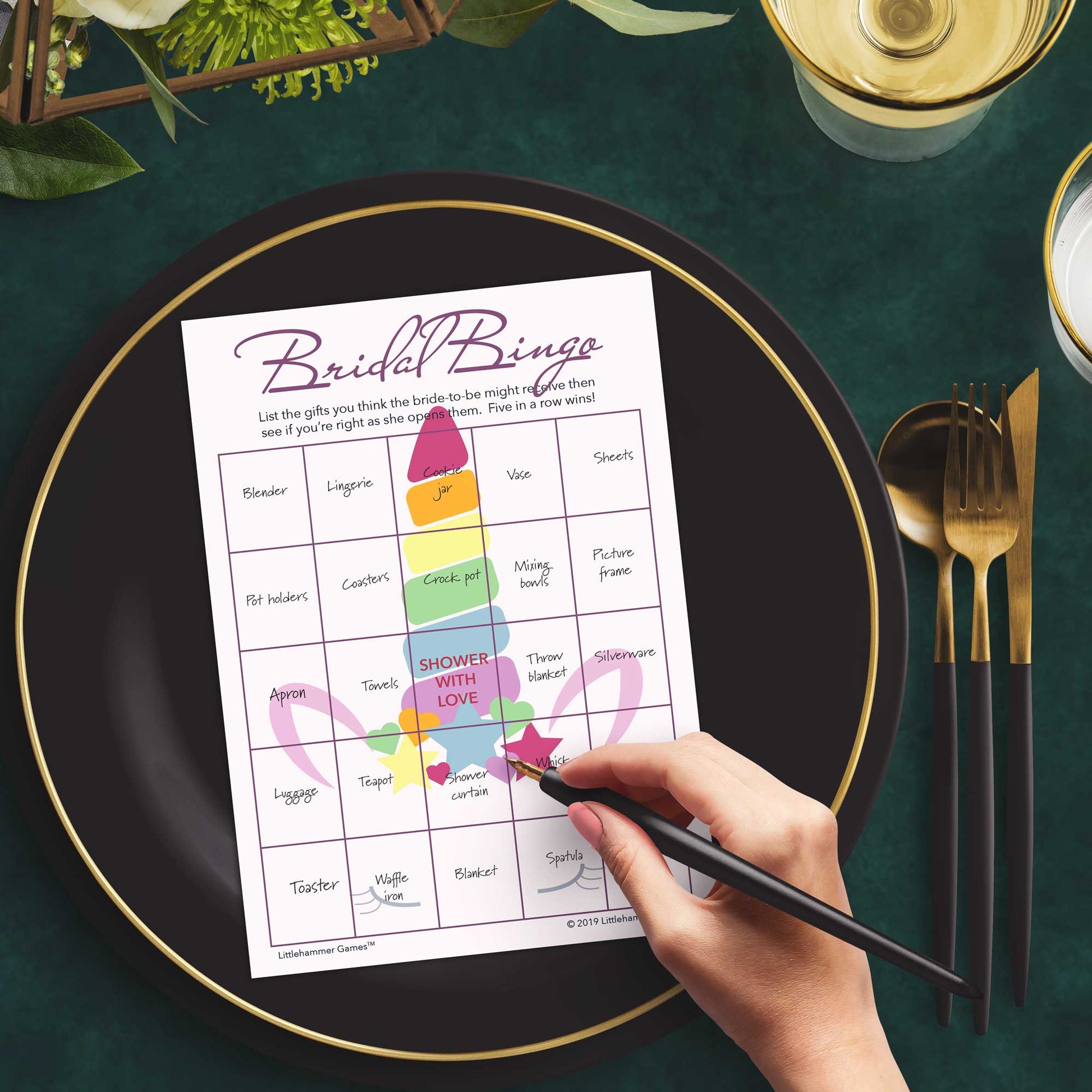Woman with a pen sitting at a dark place setting with a black and gold plate filling out a unicorn-themed Bridal Gift Bingo card