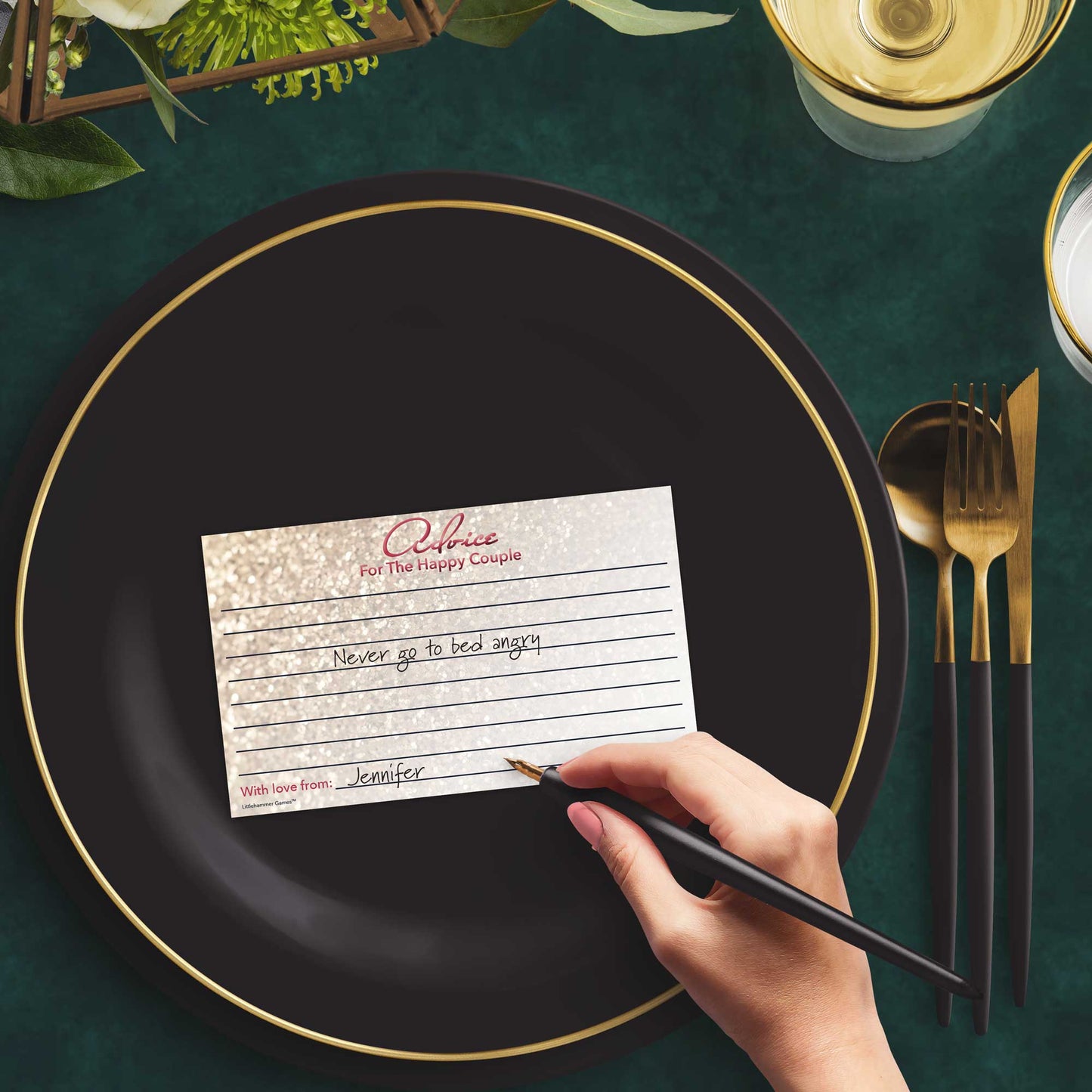 Woman with a pen sitting at a dark place setting with a black and gold plate filling out a glittery rose gold Advice for the Happy Couple card