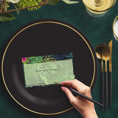 Woman with a pen sitting at a dark place setting with a black and gold plate filling out a tropical-themed Advice for the Happy Couple card