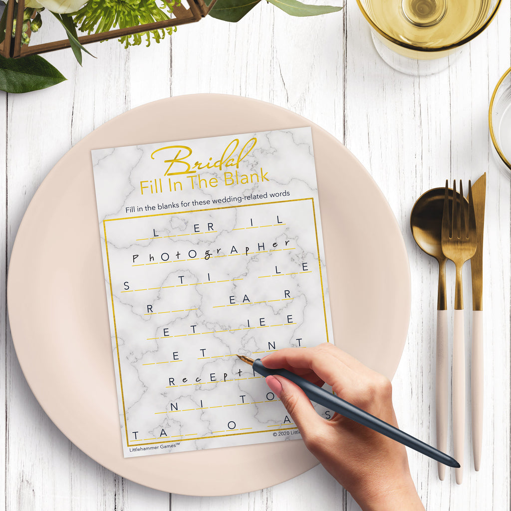 Woman’s hand holding a pen playing a gold marble Bridal Fill in the Blank game card at a light place setting