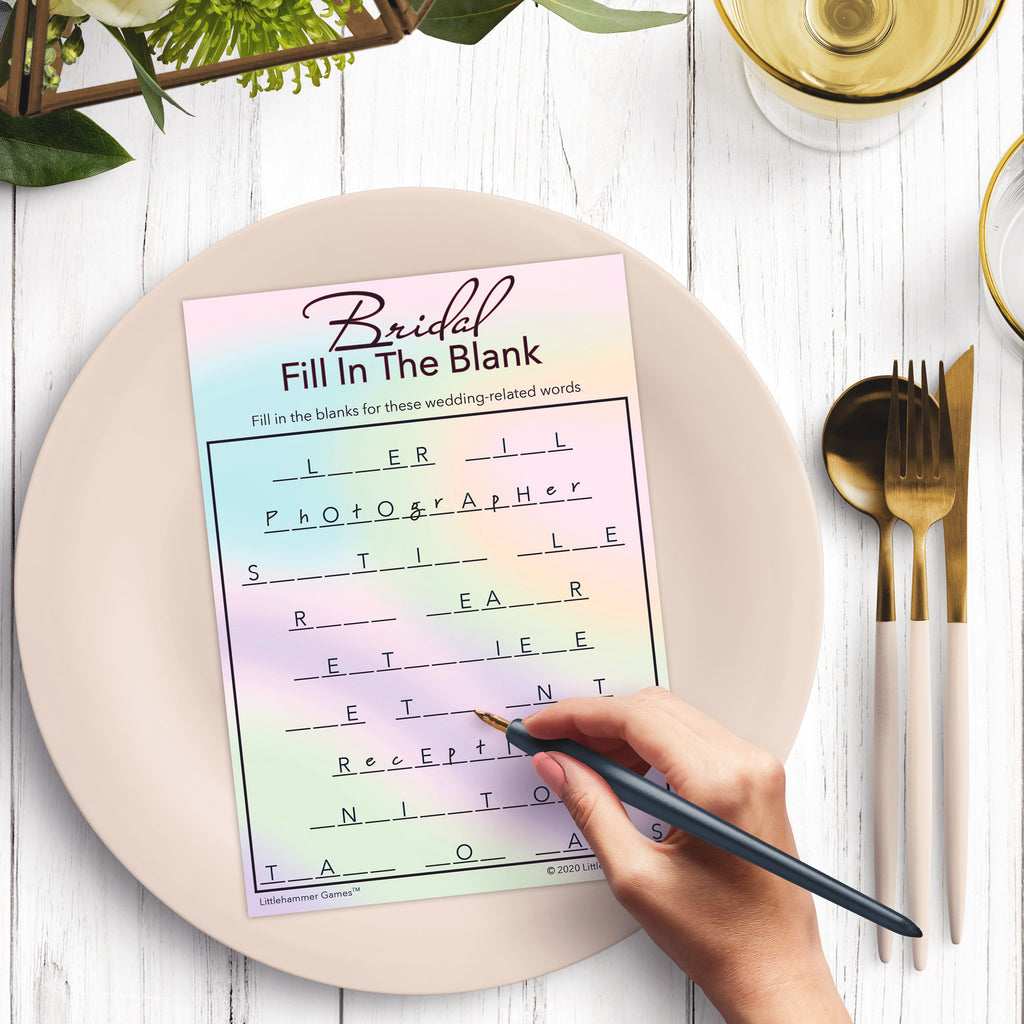 Woman’s hand holding a pen playing a hologram-themed Bridal Fill in the Blank game card at a light place setting