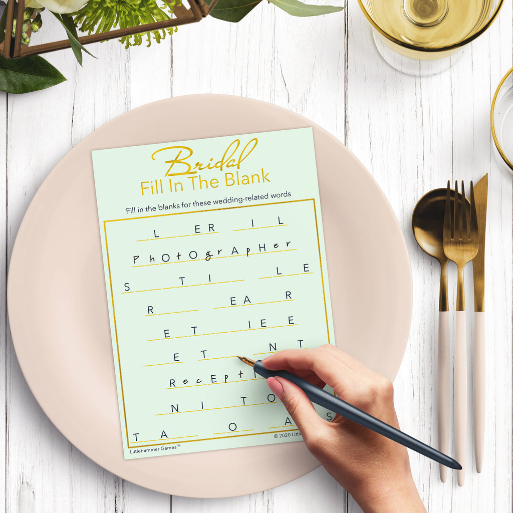 Woman’s hand holding a pen playing a mint and gold Bridal Fill in the Blank game card at a light place setting