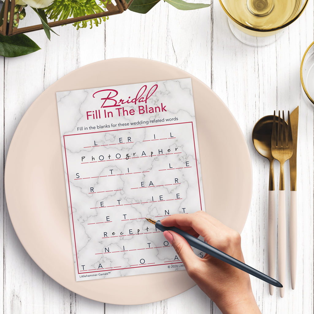 Woman’s hand holding a pen playing a rose gold marble Bridal Fill in the Blank game card at a light place setting