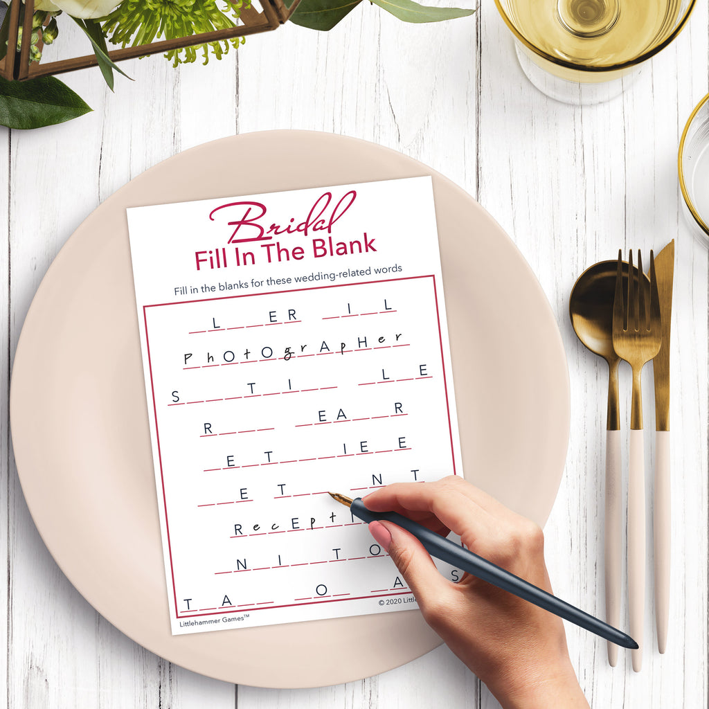 Woman’s hand holding a pen playing a white and rose gold Bridal Fill in the Blank game card at a light place setting